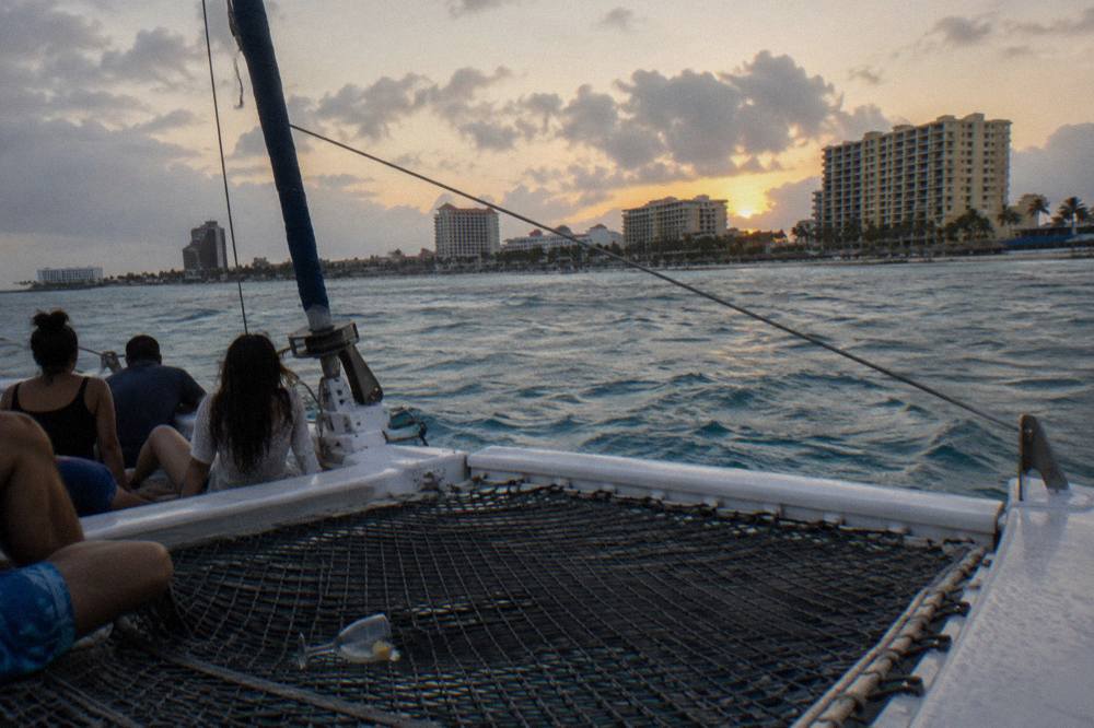 Open deck catamaran with guests seated along the trampoline nets sailing past a beachfront at dusk