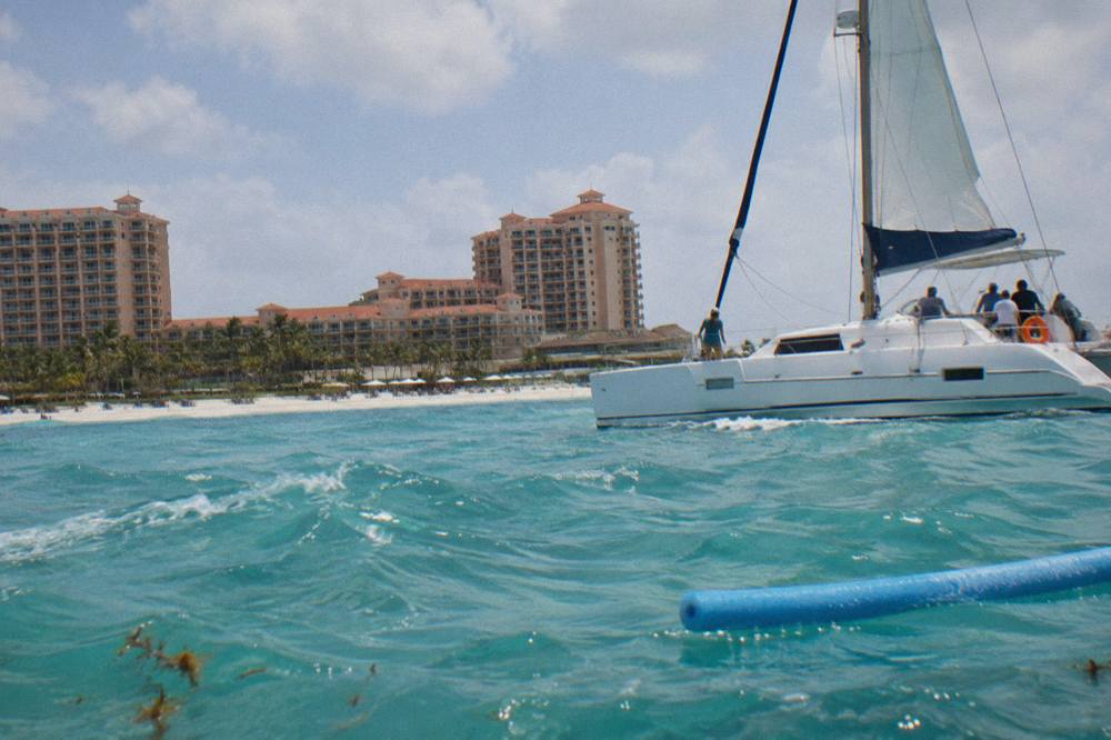 A white catamaran under sail along the calm turquoise water of Palm Beach with resort towers behind