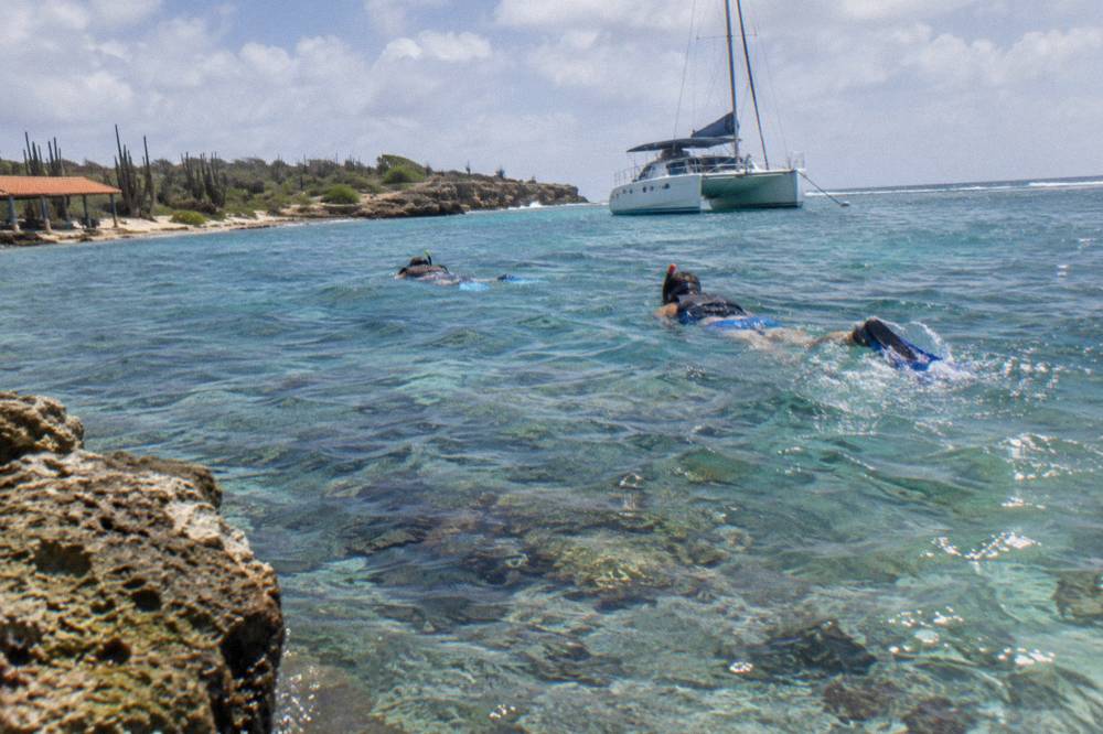 Snorkelers floating above a shallow coral reef in clear blue water near a moored catamaran
