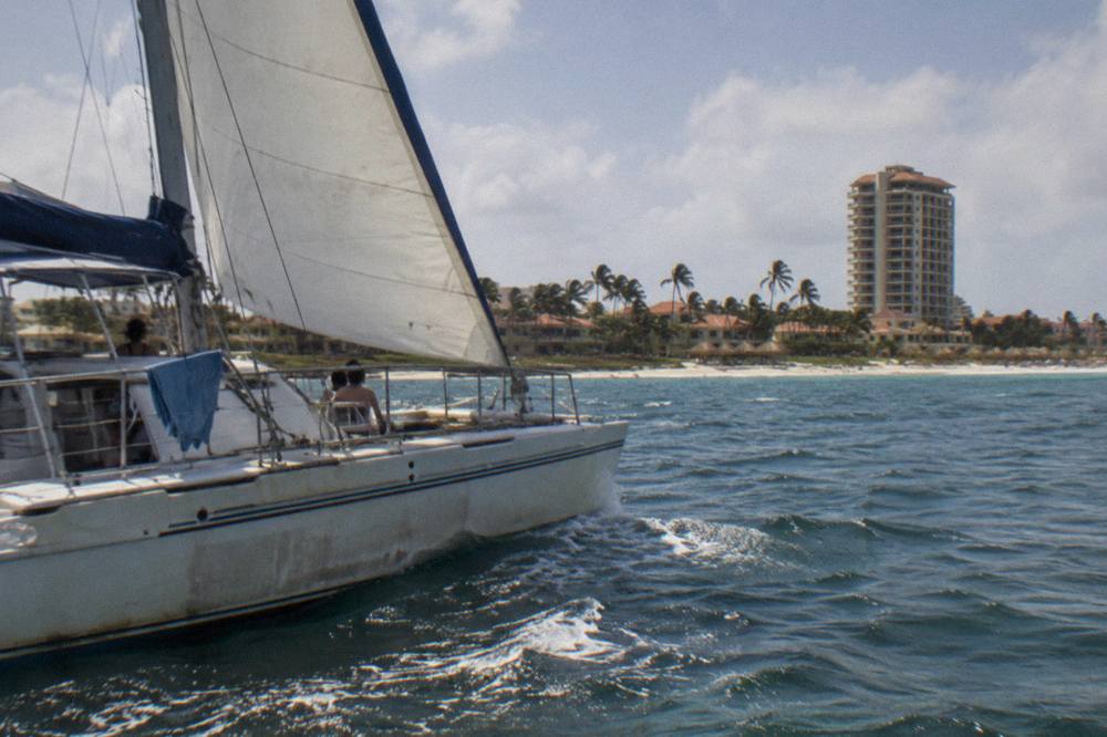 A catamaran under sail along a calm Caribbean coastline in late afternoon light
