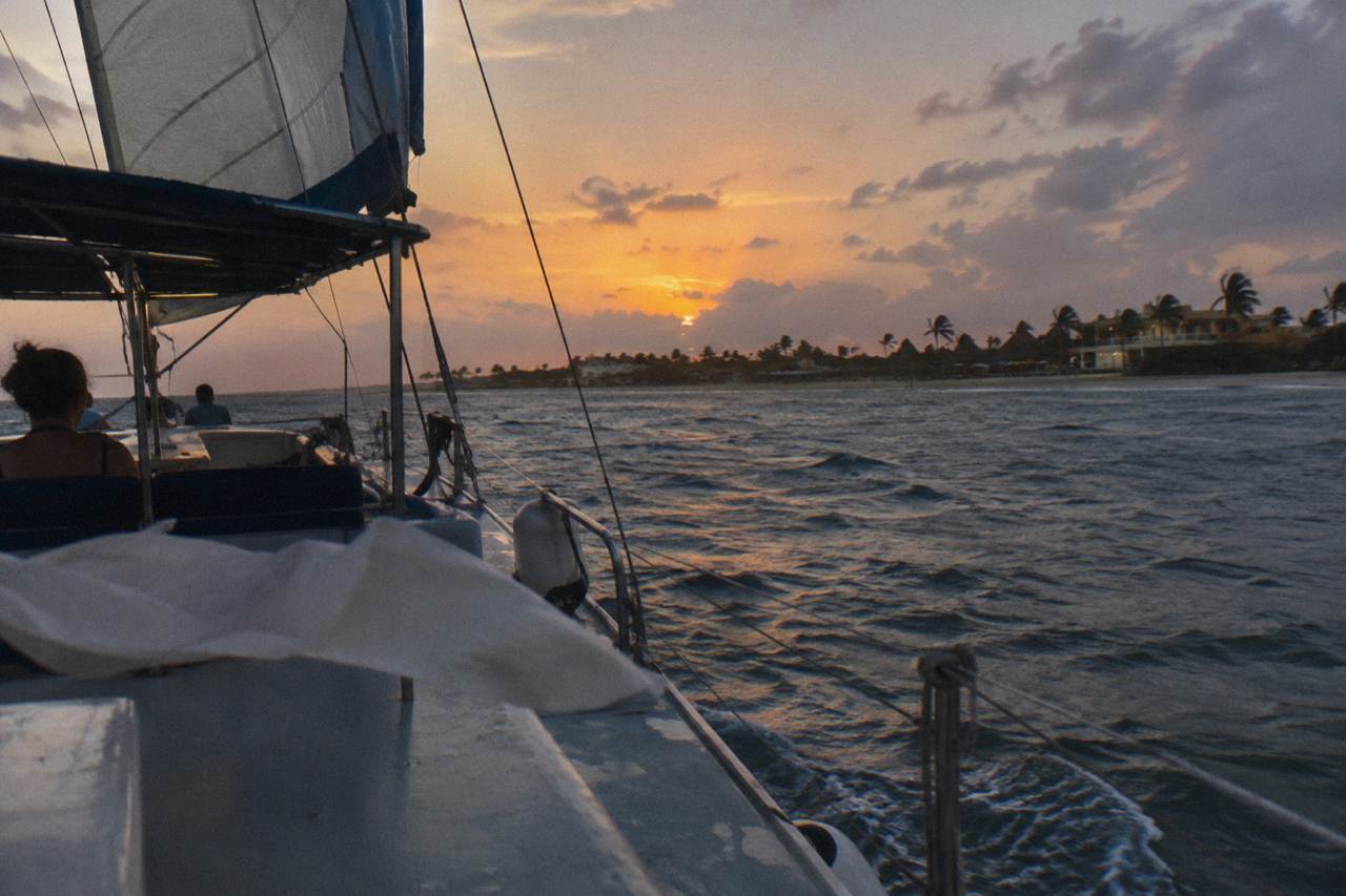 A catamaran under sail along a calm coastline at dusk with an orange sky