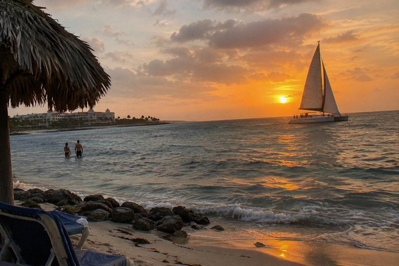 A catamaran under sail along the Aruba coast at sunset, calm sea and orange sky