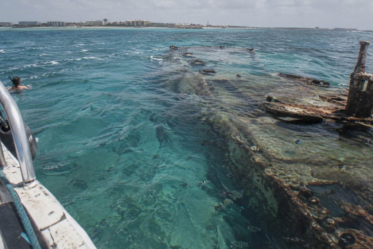 The hull of a large sunken ship lying on its side beneath clear turquoise water near the surface