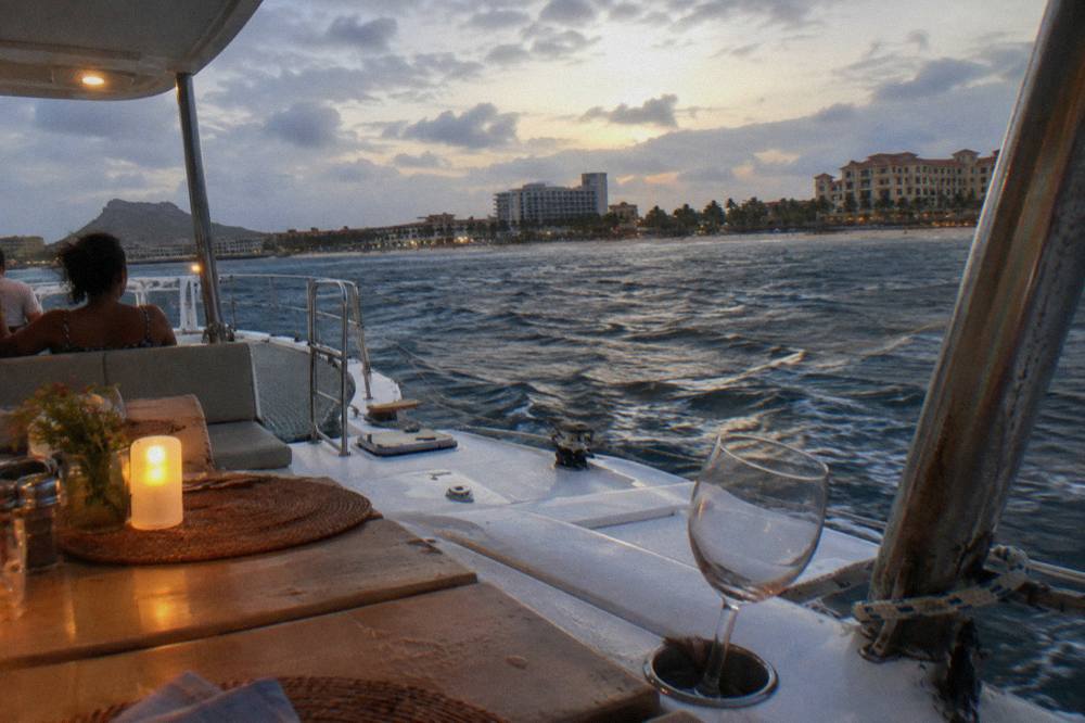 A catamaran deck at dusk with set tables and calm sea along a beach coastline
