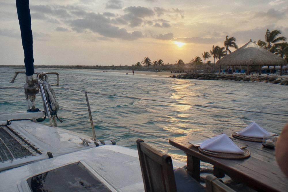 A couple's dinner table set on a catamaran deck with the sun low over calm water