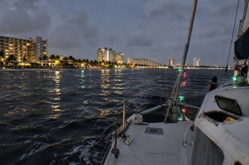 A catamaran sailing back along a beachfront at night with hotel lights reflected on calm water