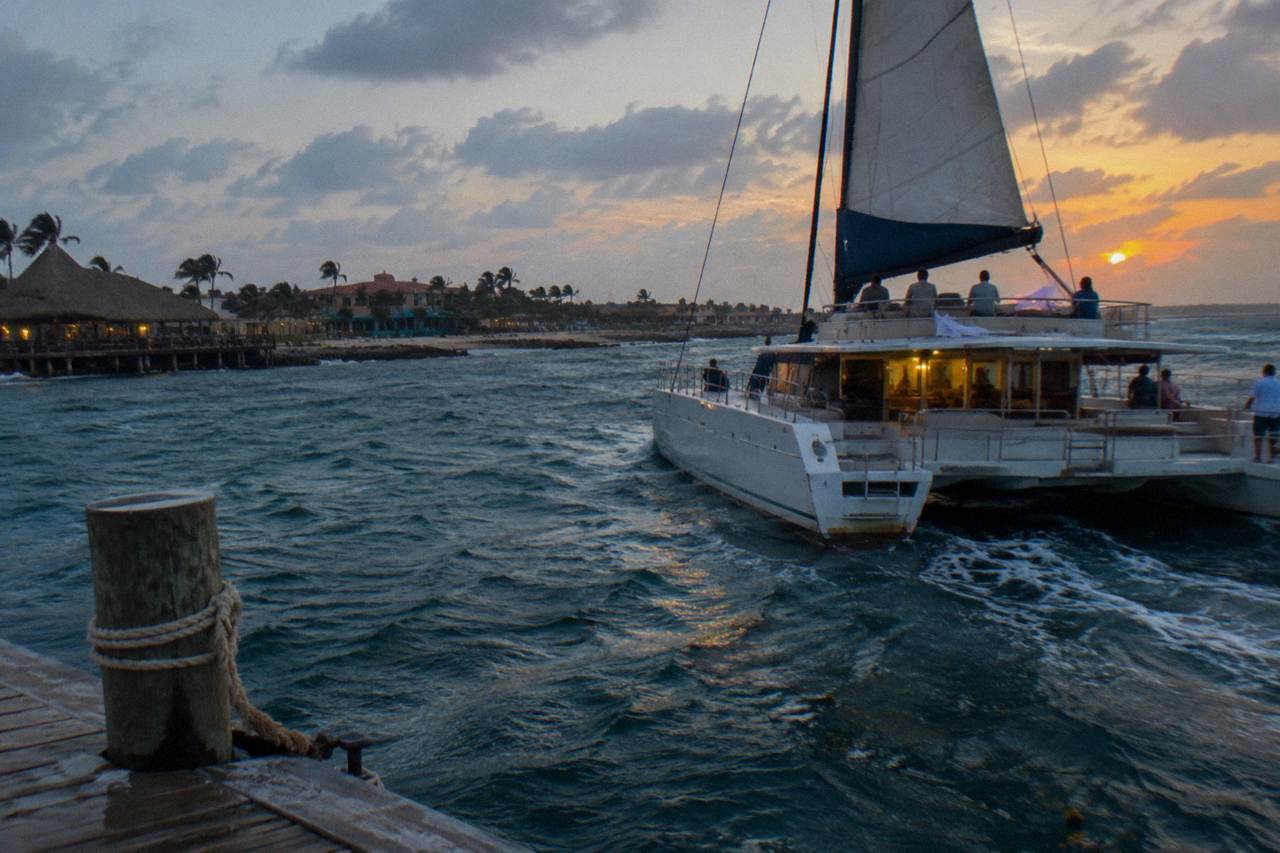 A catamaran under sail along a calm coastline at dusk with an orange sky over the water