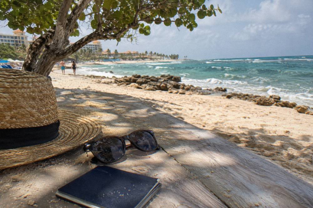 A closed passport resting on a wooden table beside a pair of sunglasses and a sun hat