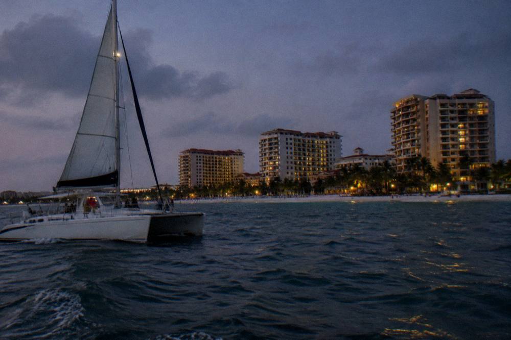 A catamaran under sail passing along a beachfront of high-rise hotels at dusk
