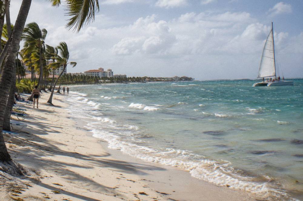 A catamaran under sail along a palm lined beach with clear shallow water