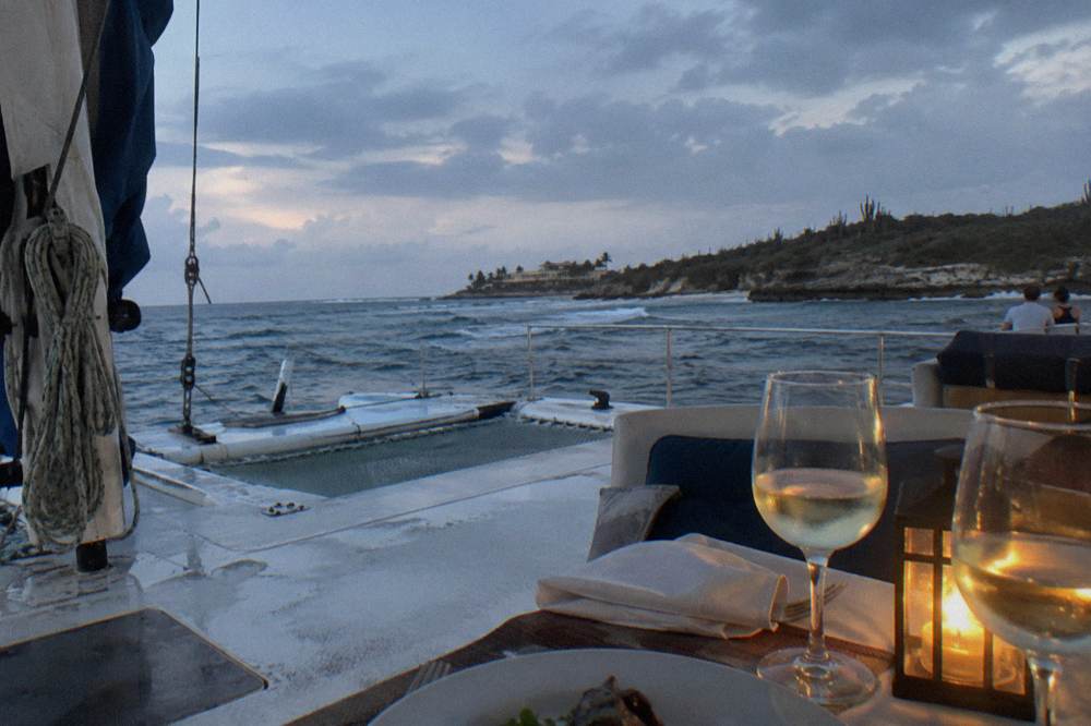 Dinner table set on a catamaran deck with wine glasses and the Aruba coastline in the background