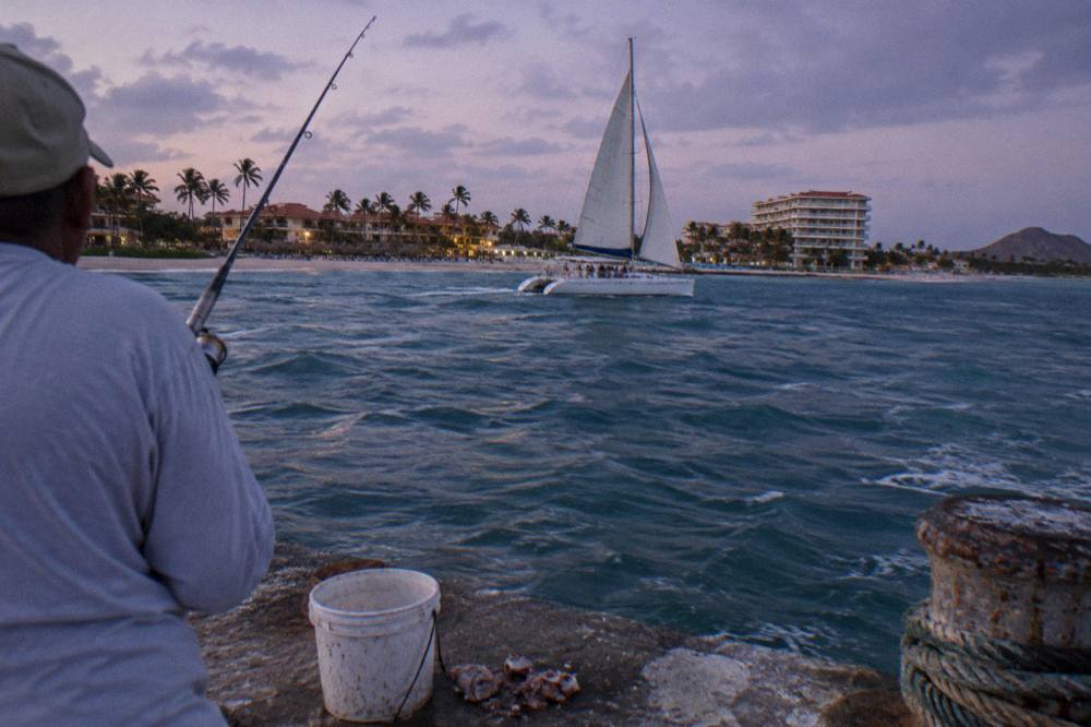 Catamaran under sail passing resorts along a beachfront at dusk