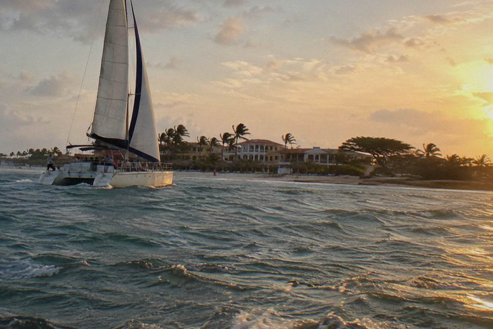 Catamaran under sail along a coastline of white sand and low-rise buildings at golden hour