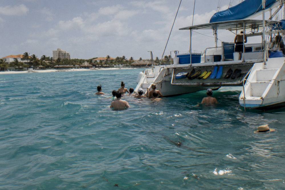 Catamaran anchored near a beach with passengers in the water beside the hull