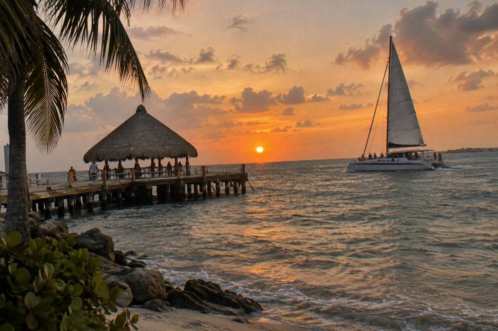 Catamaran under sail near a pier at sunset with calm sea