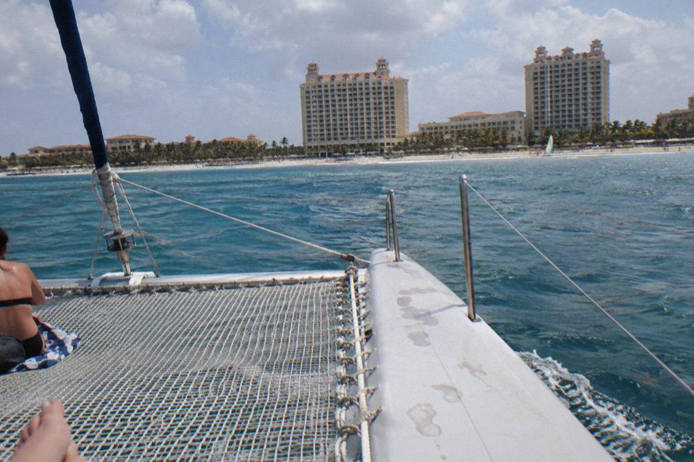 A wide catamaran deck with netting at the bow and calm blue water stretching to the Aruba shoreline