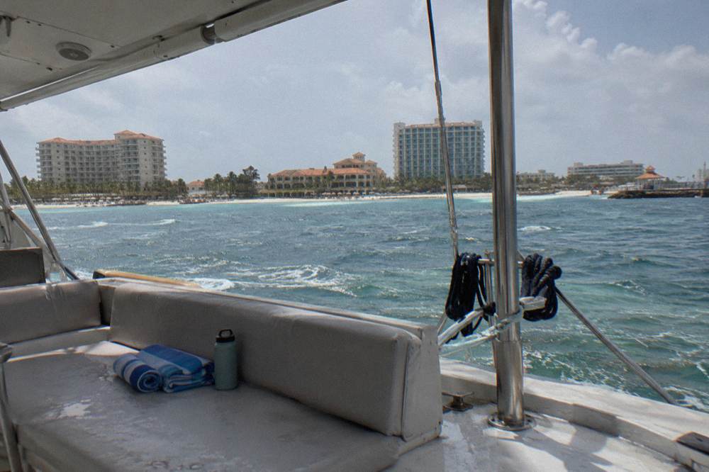 Passenger seating along the shaded aft deck of a catamaran with the Aruba beachfront on the horizon