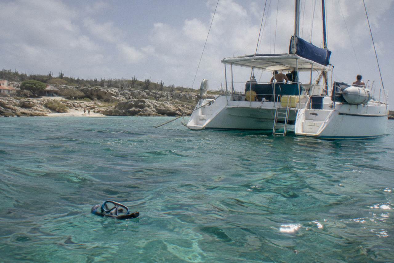 A catamaran anchored in calm turquoise water off the Aruba coast with a rocky shoreline in the distance